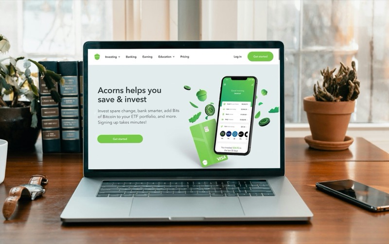 Laptop auf einem Schreibtisch zeigt die Acorns-Website mit einer mobilen App und Kreditkarten.