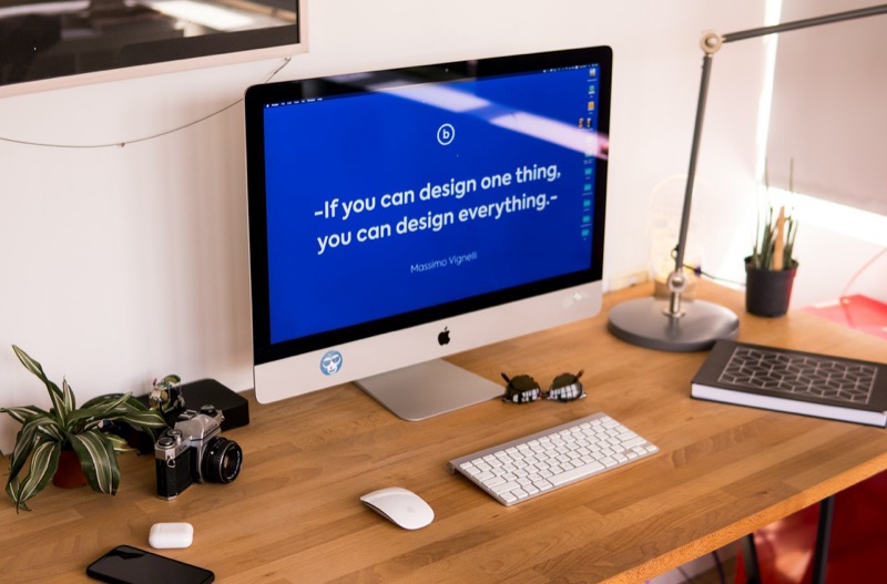 Ein Schreibtisch mit iMac, Tastatur, Maus, Brille, Notizbuch, Stiften, Lampe und einer Pflanze.