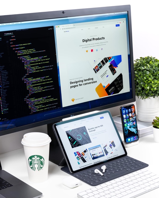 Ein Schreibtisch mit Monitor, der Code und eine Landingpage anzeigt, einem Tablet, einem Smartphone und einem Starbucks-Becher.