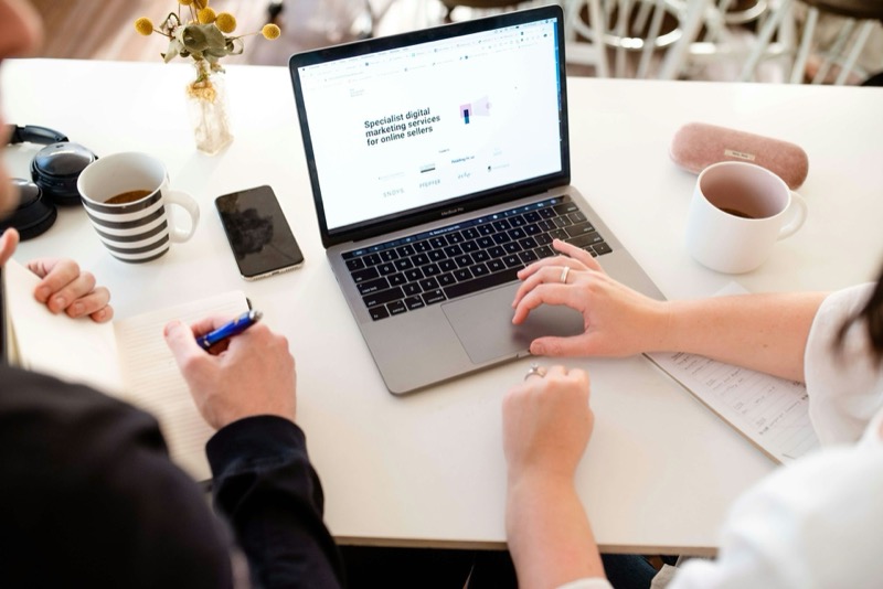 Zwei Personen arbeiten an einem Laptop auf einem weißen Tisch, mit Kaffeebechern, Kopfhörern und einem Smartphone.