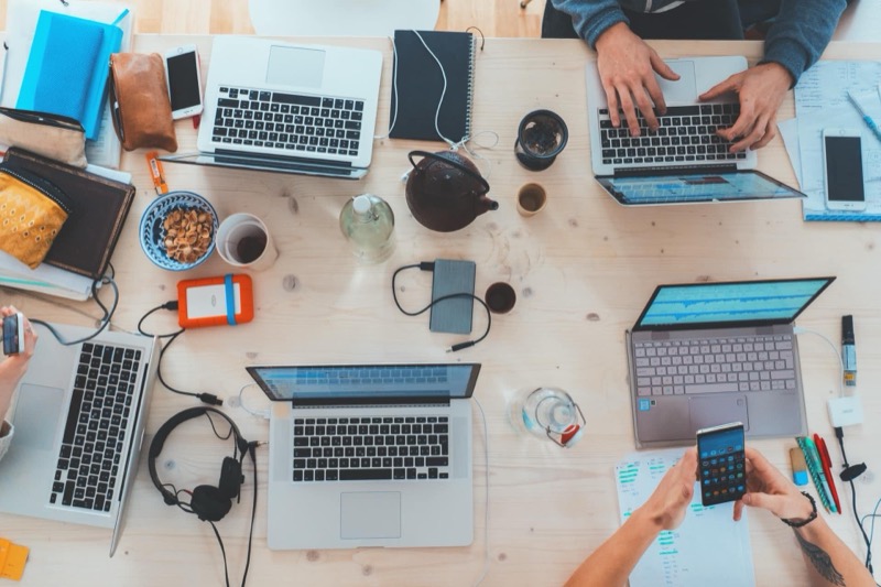 Blick von oben auf vier Personen, die an einem Holztisch mit Laptops, Smartphones, Kopfhörern und Notizbüchern arbeiten.