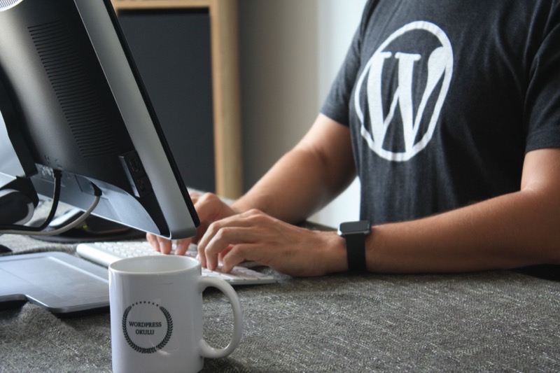 Mann im WordPress-T-Shirt tippt an einem Computer, eine Tasse mit „WordPress-Disco“-Logo steht auf dem Schreibtisch.